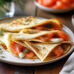Crêpes au saumon fumé garnies de crème citronnée à l'aneth, pliées en quatre et dressées sur une assiette blanche avec des tranches de saumon rose et un quartier de citron