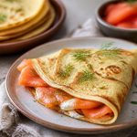Crêpes au saumon fumé garnies de crème citronnée à l'aneth et de zestes de citron, servies pliées en quatre sur une assiette blanche