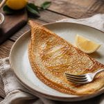 Recette de crêpes pour 1 personne : assiette de crêpes dorées et moelleuses avec un filet de citron et du sucre, sur une table en bois clair